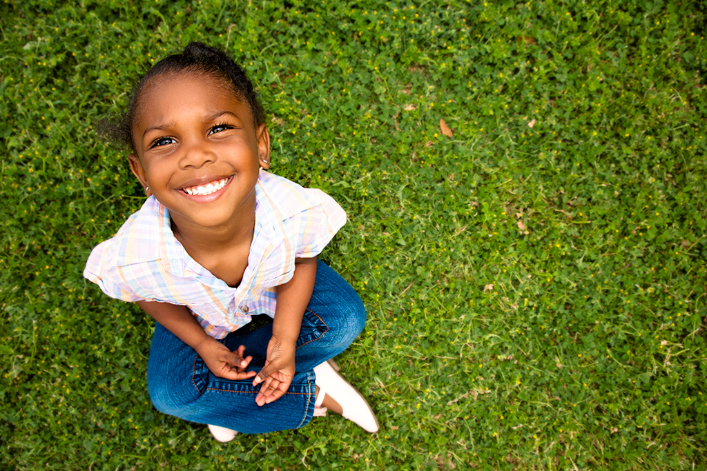 A young African-American girl is sitting in the grass, smiling.